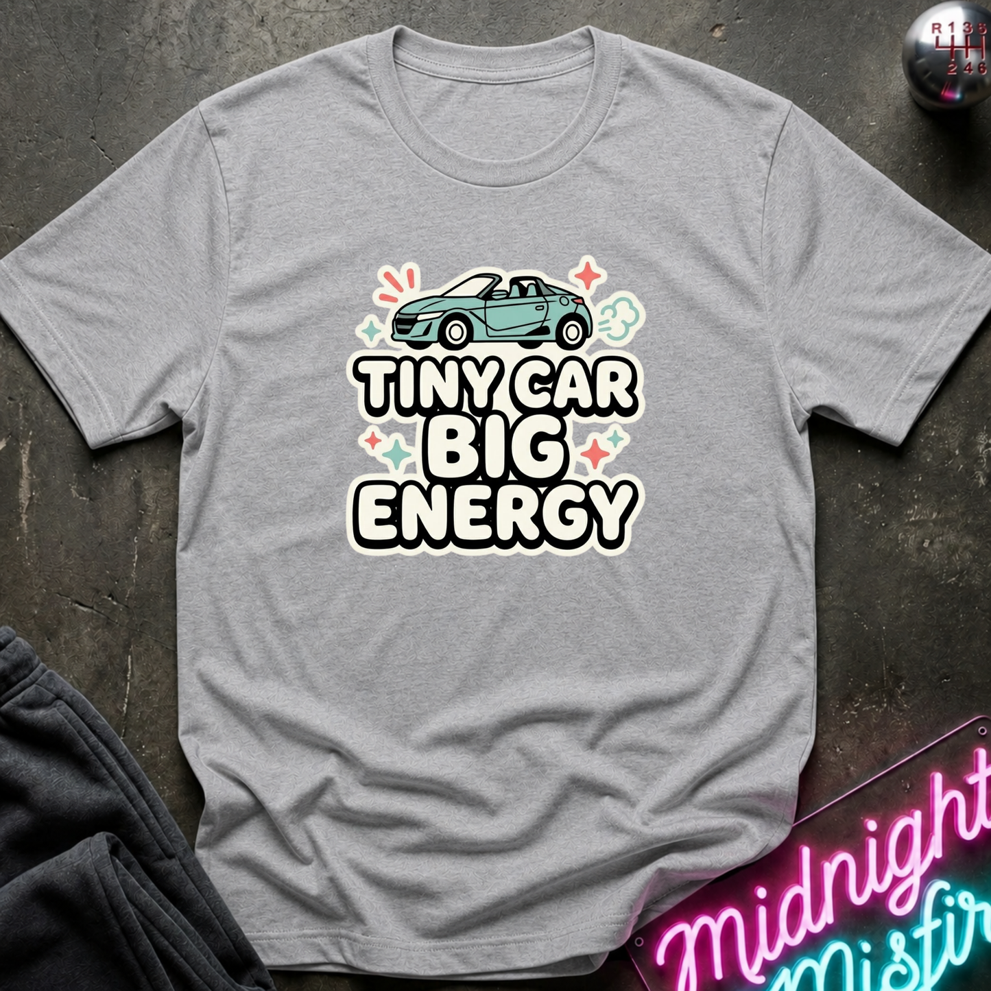 A heather gray short-sleeve t-shirt with a graphic print of a small teal convertible car and the text 'TINY CAR BIG ENERGY' in white with black outlines, surrounded by small colorful star and spark motifs. The shirt is laid flat on a dark textured surface with a neon sign and dark pants partially visible as props.