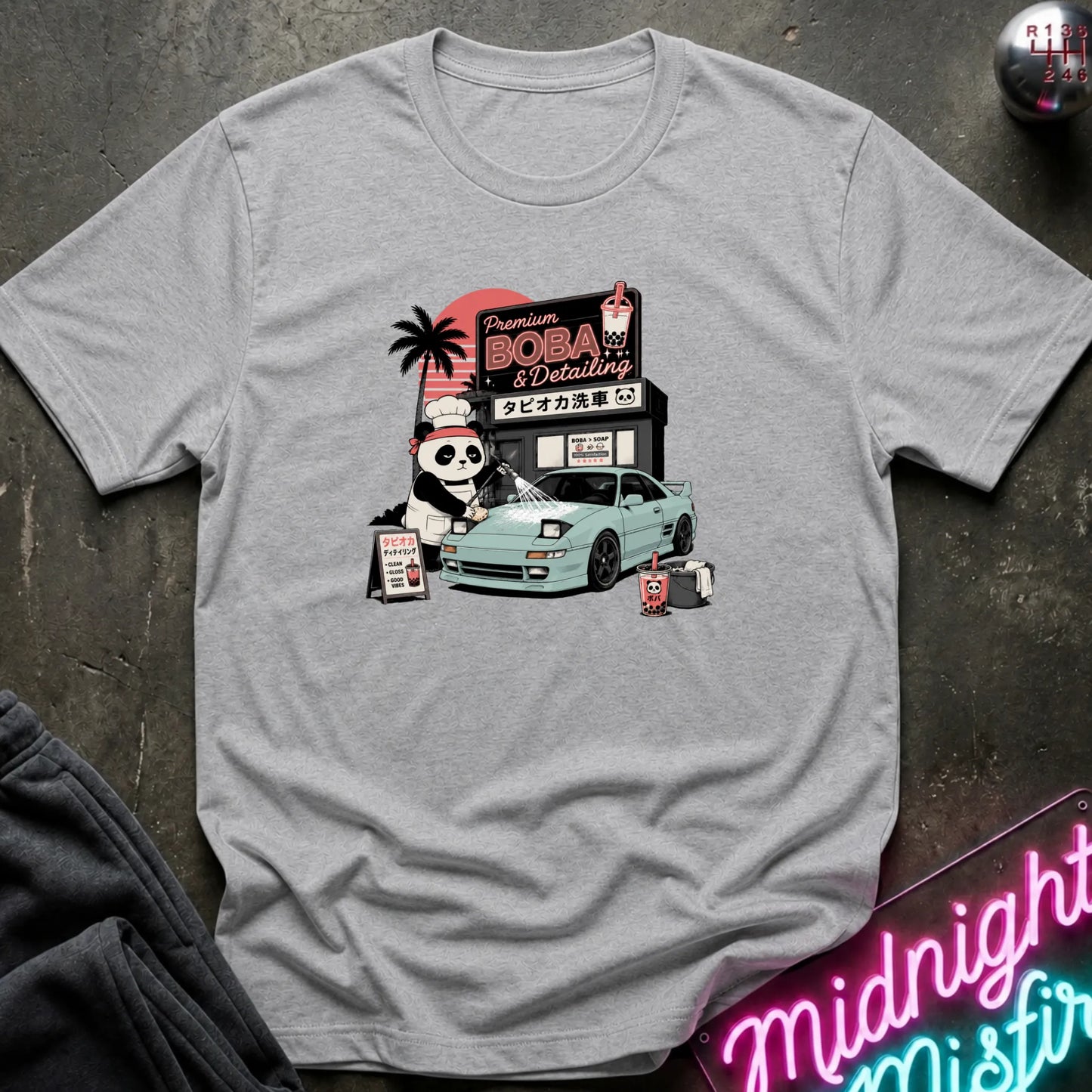 A light gray short-sleeve graphic T-shirt laid flat on a dark textured surface. The shirt features a colorful retro-style print of a panda character, a light blue sports car, and a sign reading 'Premium BOBA & Coffee' with Japanese text.