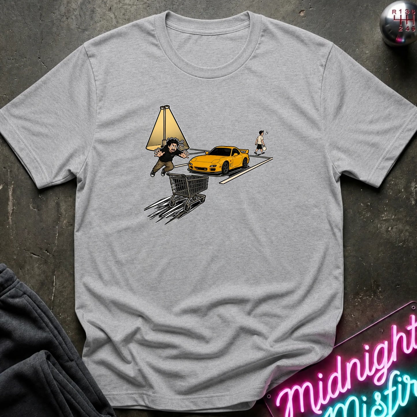 A light gray heather crewneck t-shirt laid flat on a dark textured surface. The shirt features a graphic print of a yellow sports car, a shopping cart and a person walking, styled with a neon sign and dark pants in the background.