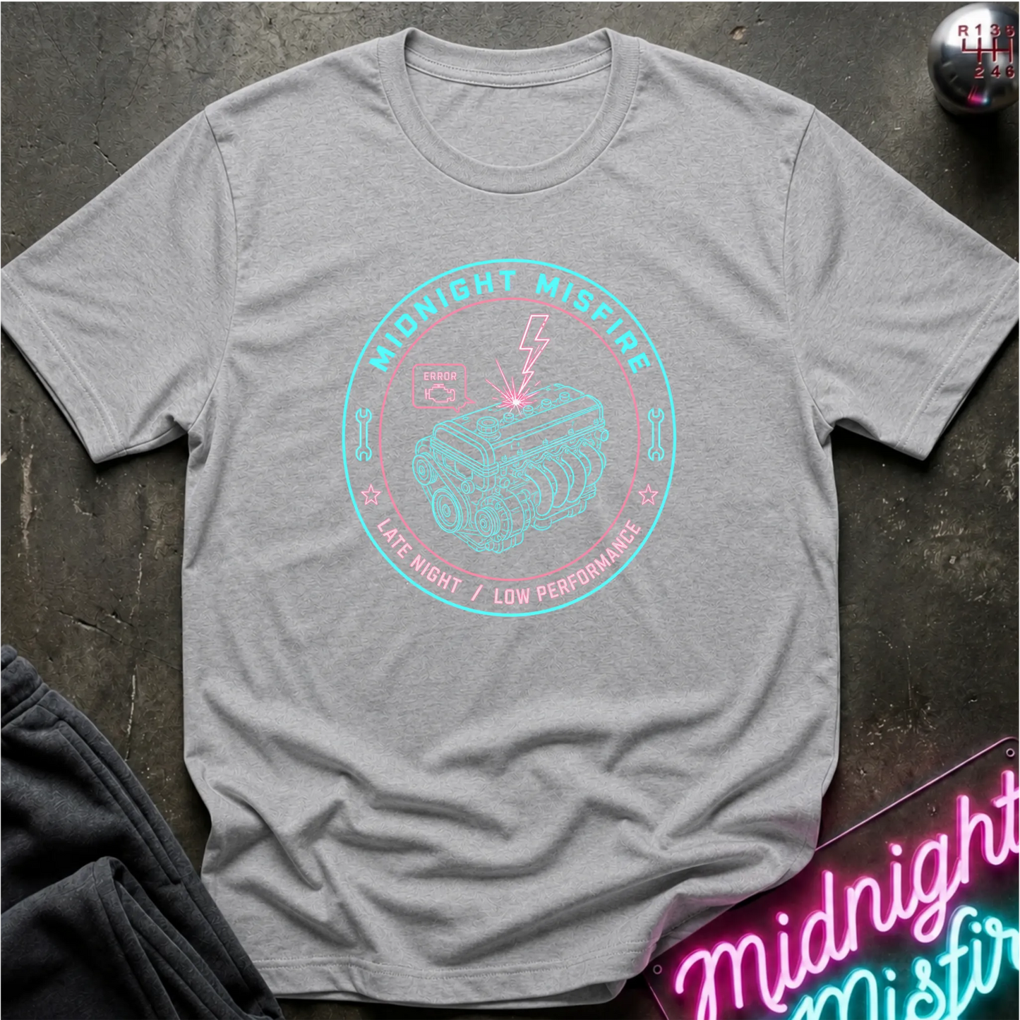 A heather gray short-sleeve t-shirt laid flat on a dark textured surface, featuring a circular graphic print in light blue and pink with an engine illustration and the words 'Midnight Mispire' and 'Late Night / Low Performance.'.