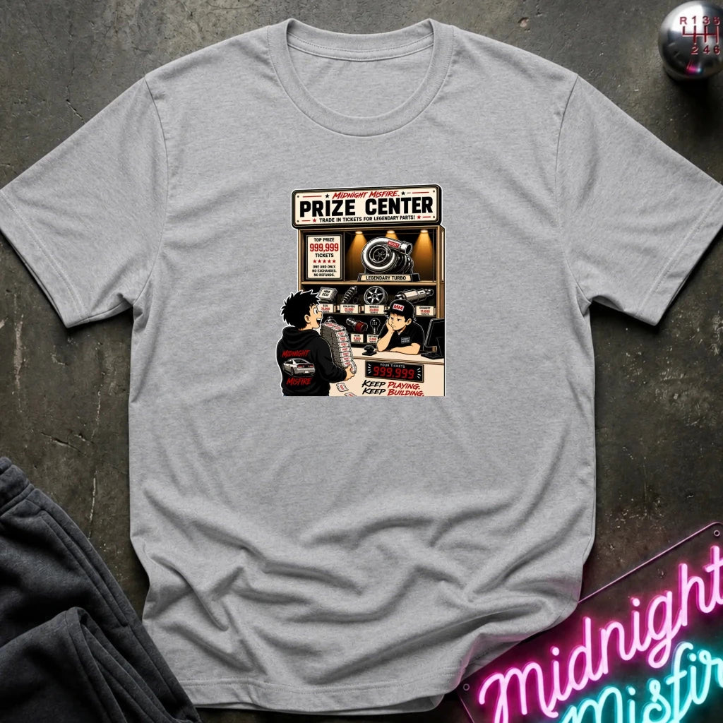 A light gray  short-sleeve crewneck t-shirt laid flat on a dark textured surface, featuring a vintage-style graphic print of a 'Prize Center' with illustrated characters and neon signs in the background.
