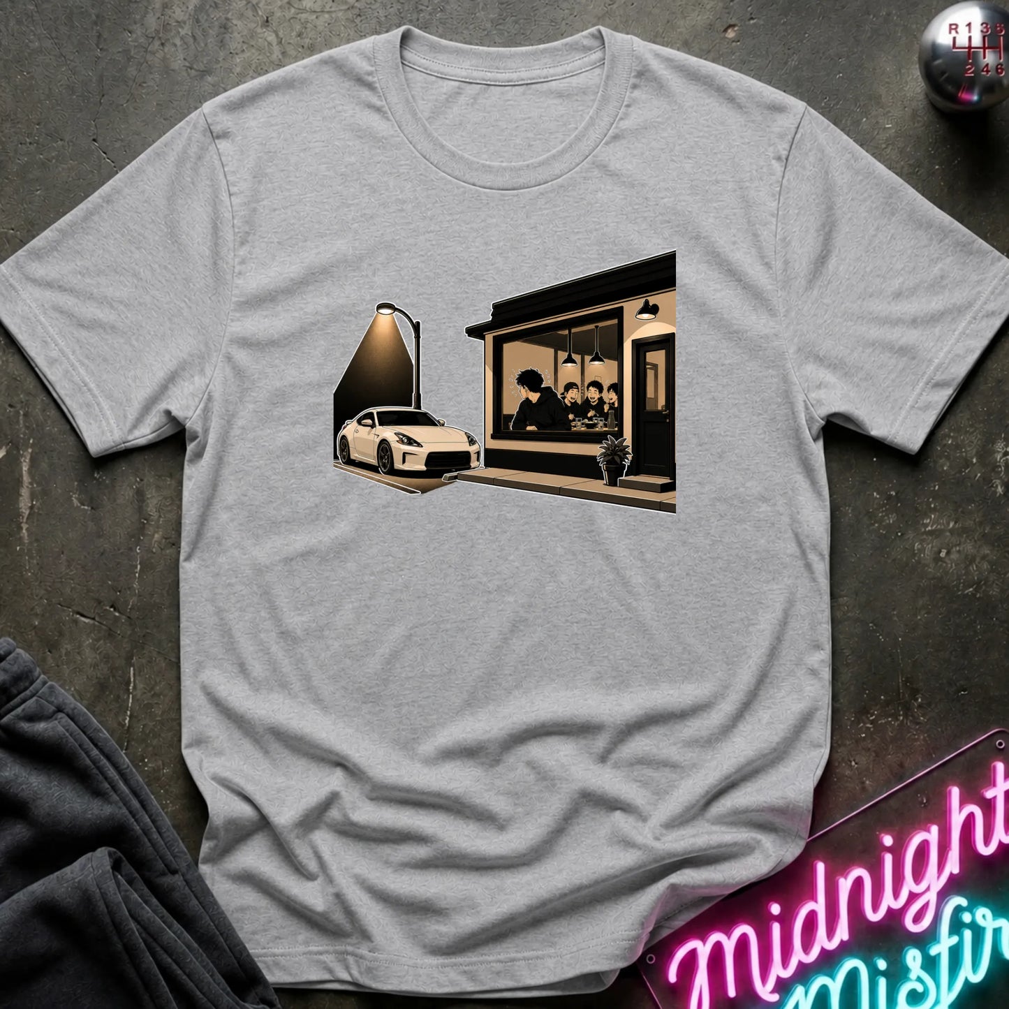 A light gray heather crewneck t-shirt featuring a graphic print of a white sports car parked outside a cafe at night, styled flat-lay on a dark textured surface with a neon sign and dark pants partially visible.