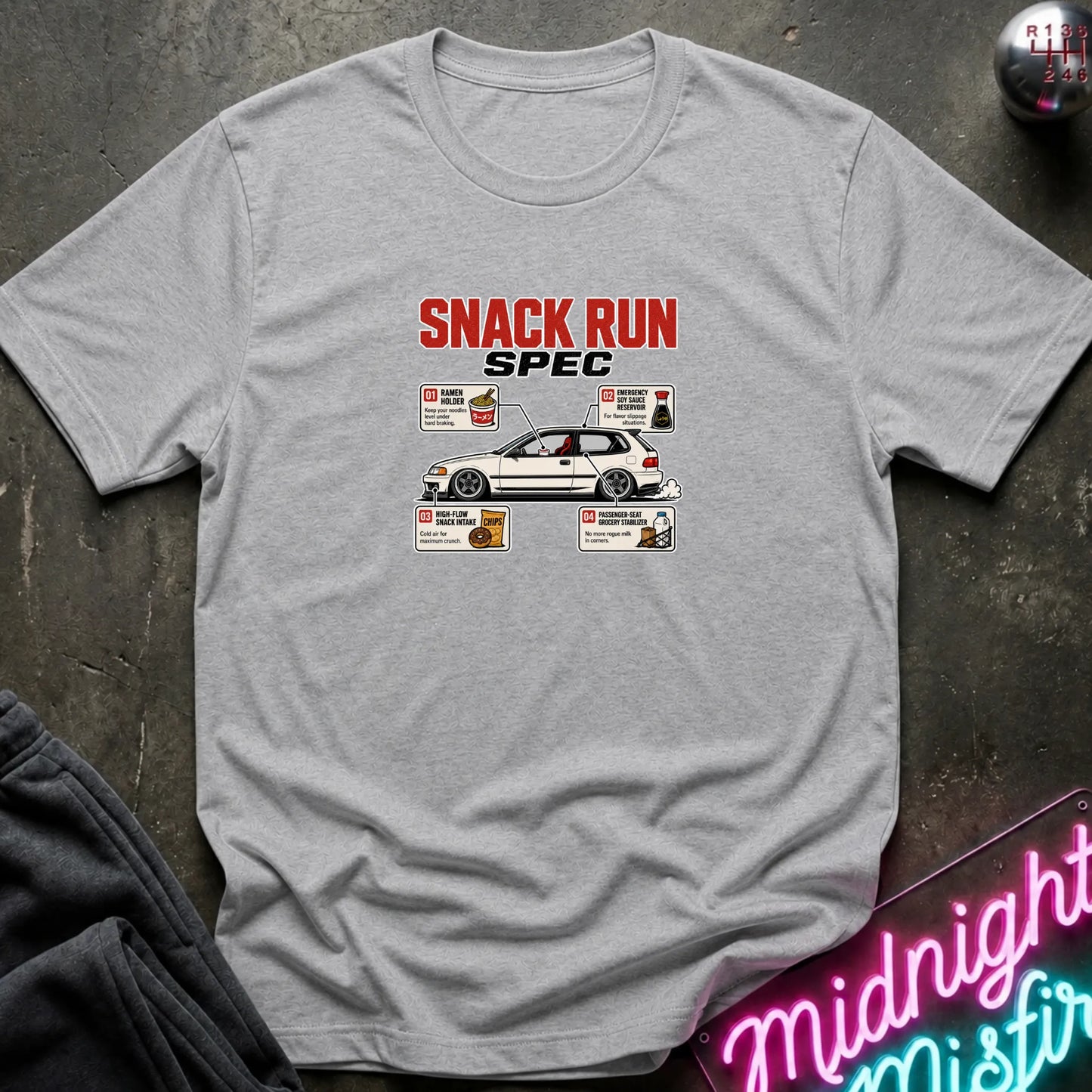 A light gray heather crewneck t-shirt featuring a 'SNACK RUN SPEC' graphic print on the chest. The graphic includes a white hatchback car and various food illustrations styled like menu items.