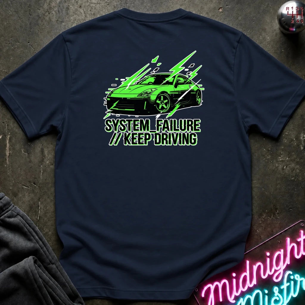 navy short-sleeve t-shirt laid flat on a dark textured surface, featuring a graphic of a green sports car with text 'SYSTEM FAILURE // KEEP DRIVING' in green. A neon sign and dark clothing are partially visible as props.