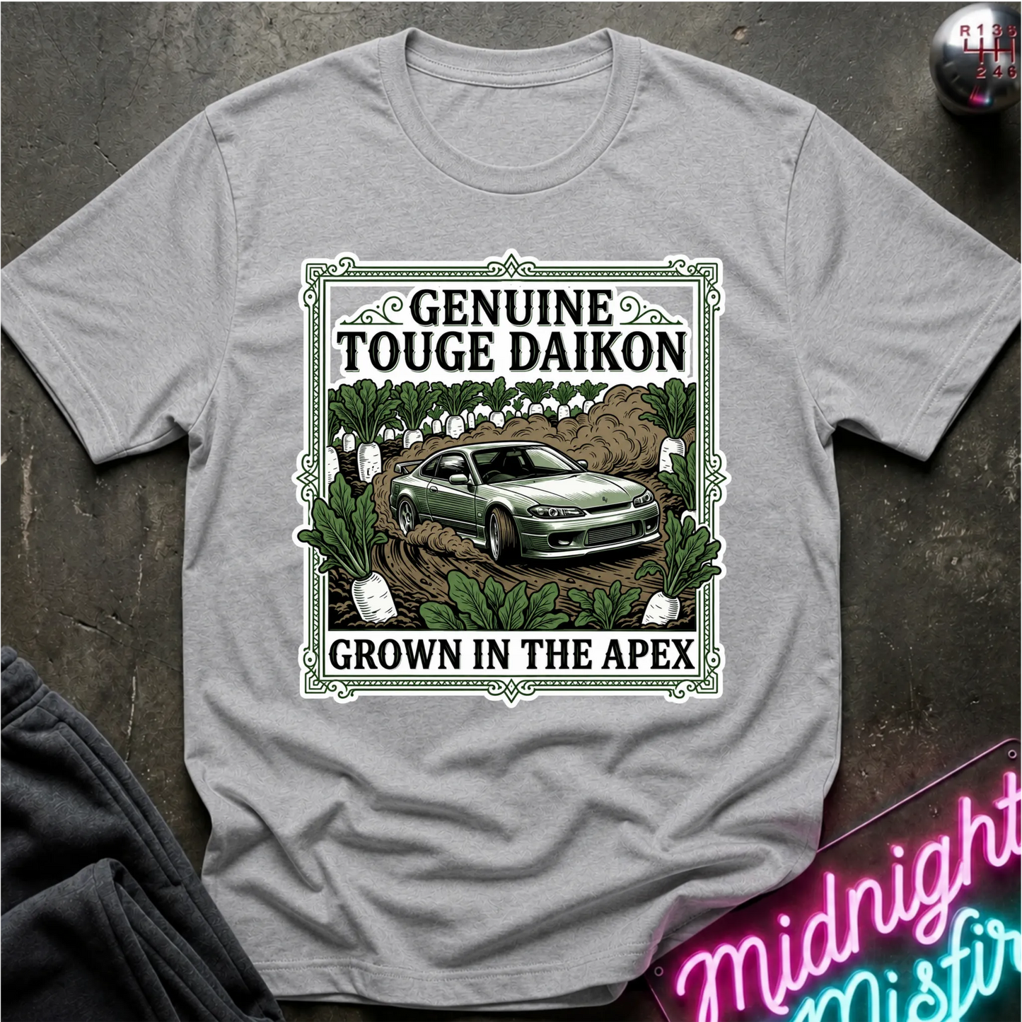 A light gray short-sleeve graphic T-shirt laid flat on a dark textured surface. The shirt features a large rectangular front print with the words 'GENUINE TOUGHE DAIKON' and 'GROWN IN THE APEX'.