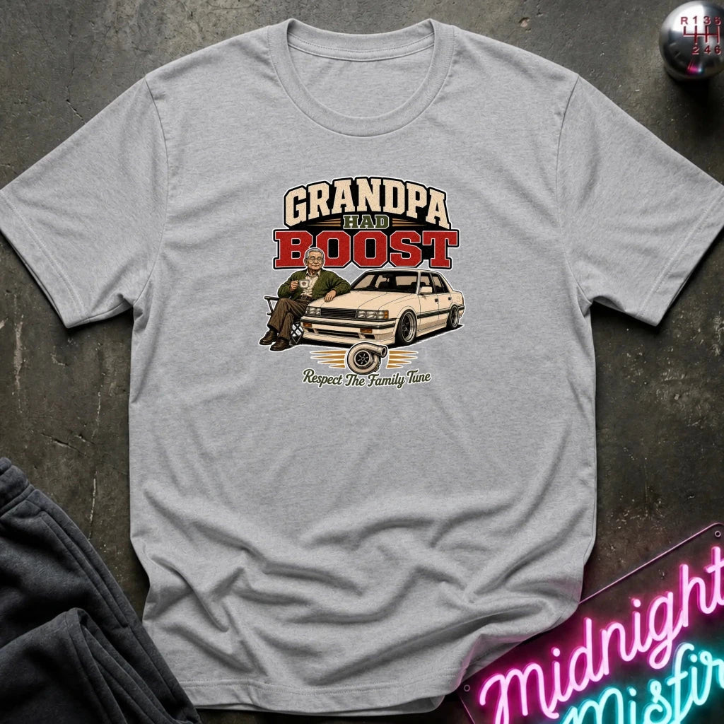 grey short-sleeve graphic t-shirt laid flat on a dark textured surface. The graphic features the text 'GRANDPA RAW BOOST' in beige and red, above an illustration of a man sitting in a car with the phrase 'Respect The Family Tune' below.