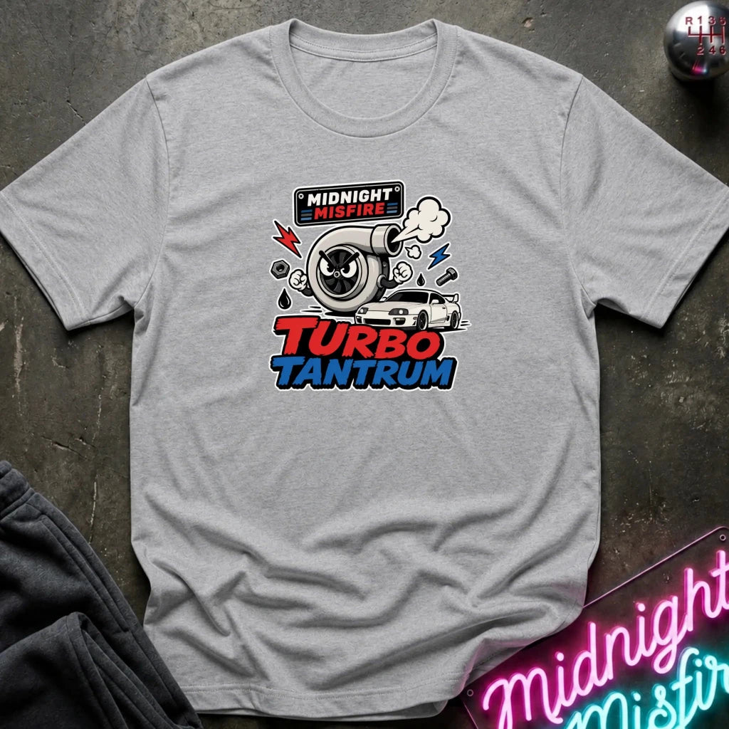 A light heather gray short-sleeve t-shirt with a 'Midnight Misfire Turbo Tantrum' graphic on the chest, displayed flat on a dark textured surface with a neon sign and dark pants in the background.