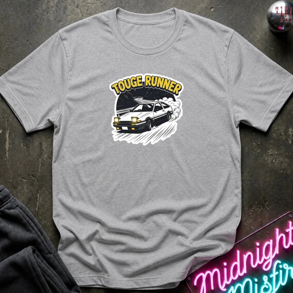 A heather gray short-sleeve t-shirt laid flat on a dark textured surface, featuring a graphic of a black sports car with the words 'TOUGE RUNNER' in yellow above it. A neon sign and folded dark fabric are partially visible around the edges.