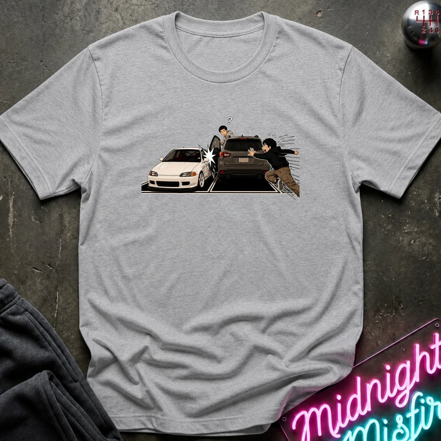 A light gray crewneck t-shirt featuring a graphic print of a white sports car and a dark SUV with characters laid flat on a dark textured surface.