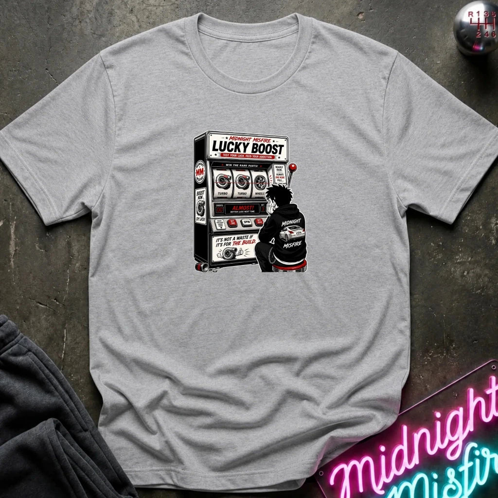 A light gray crewneck t-shirt laid flat on a dark textured surface, featuring a graphic print of a person sitting in front of a slot machine with the text 'LUCKY BOOST'.