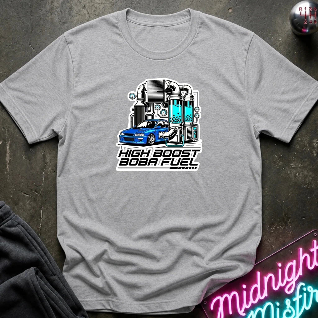 A heather gray short-sleeve t-shirt with a graphic of a blue sports car and a high-speed turbocharger, with the text 'HIGH BOOST BOBA FUEL' underneath. The shirt is displayed flat on a dark textured surface with a neon sign and dark pants partially visible.