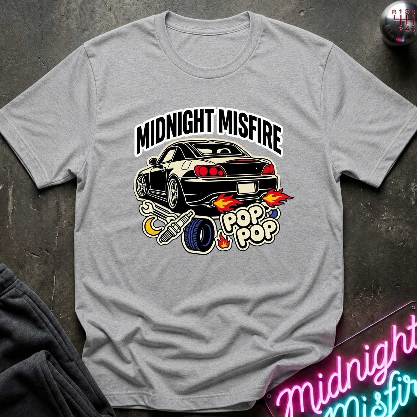 A light gray heather crewneck t-shirt featuring a graphic of a black sports car with flames and the text 'MIDNIGHT MISFIRE' and 'POP POP'. The shirt is styled flat on a dark textured surface with a neon sign and dark pants partially visible.