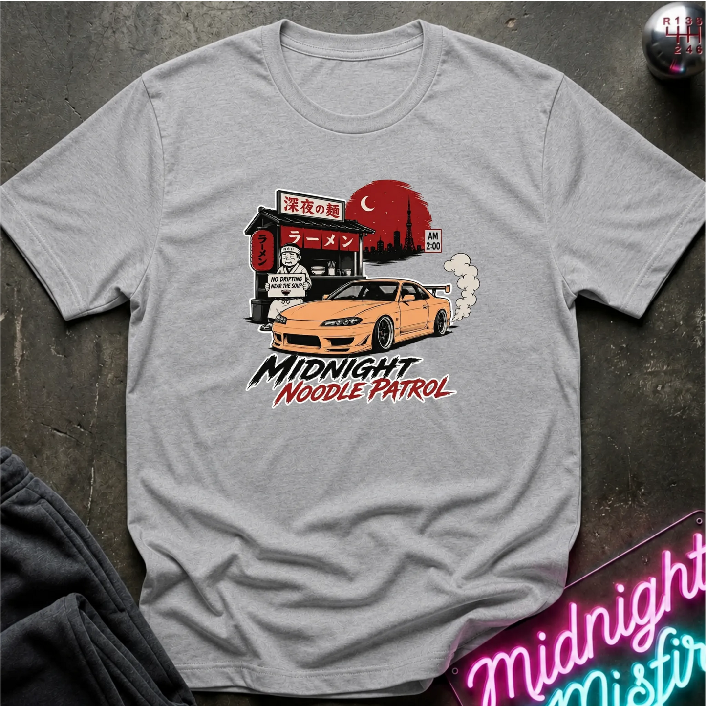 A light gray heather crewneck t-shirt featuring a graphic print of a yellow sports car in front of a ramen shop with a red full moon, and the text 'MIDNIGHT NOODLE PATROL'.