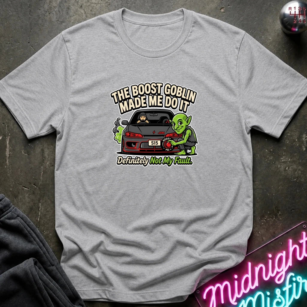A light gray heather short-sleeve graphic t-shirt laid flat on a dark textured surface. The graphic features a green cartoon character next to a red sports car with the text 'THE BOOST GOBLIN MADE ME DO IT' above and 'Definitely Not My Fault.