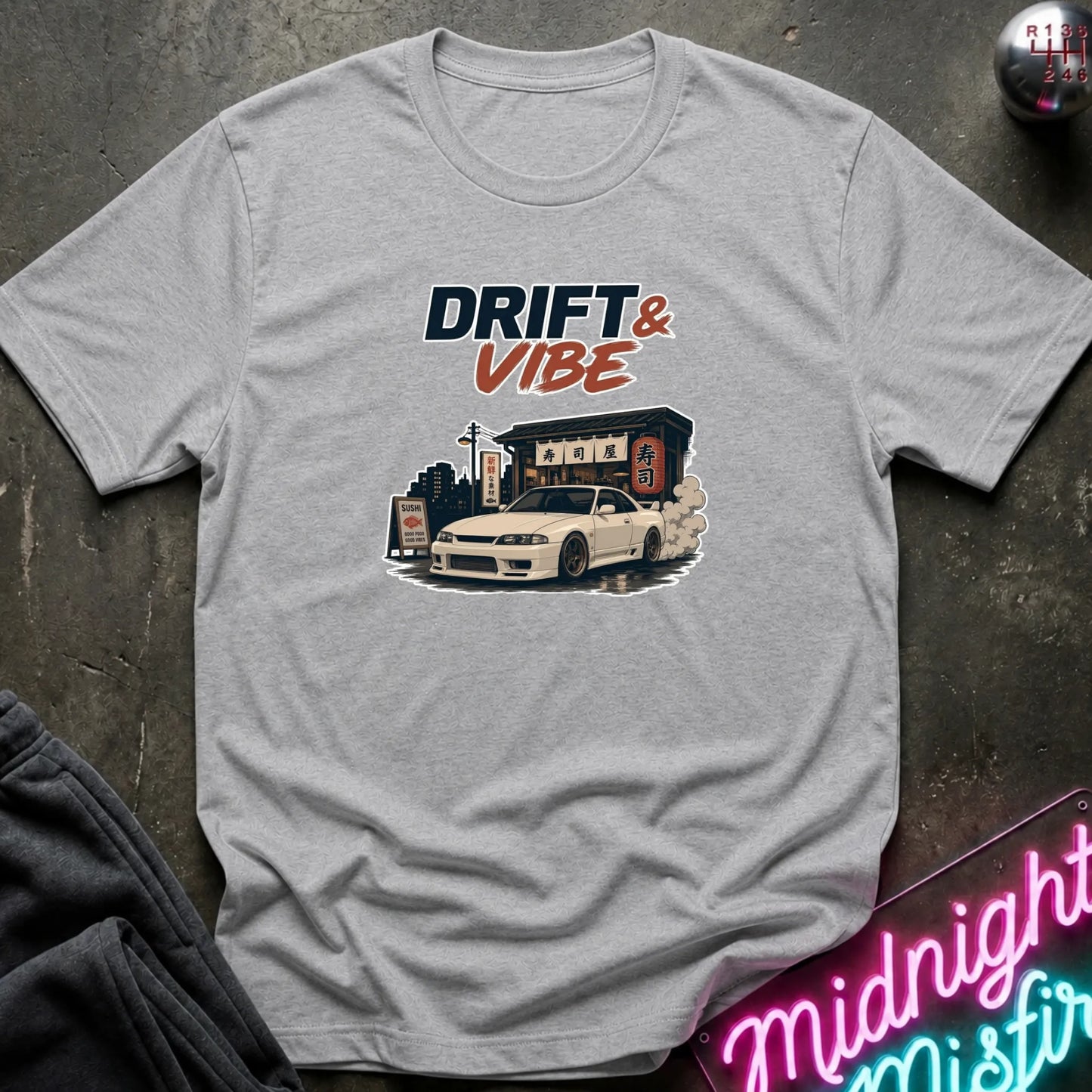 A light heather gray t-shirt laid flat on a dark textured surface, featuring a graphic print of a white sports car in front of a building with the text 'DRIFT & VIBE'.