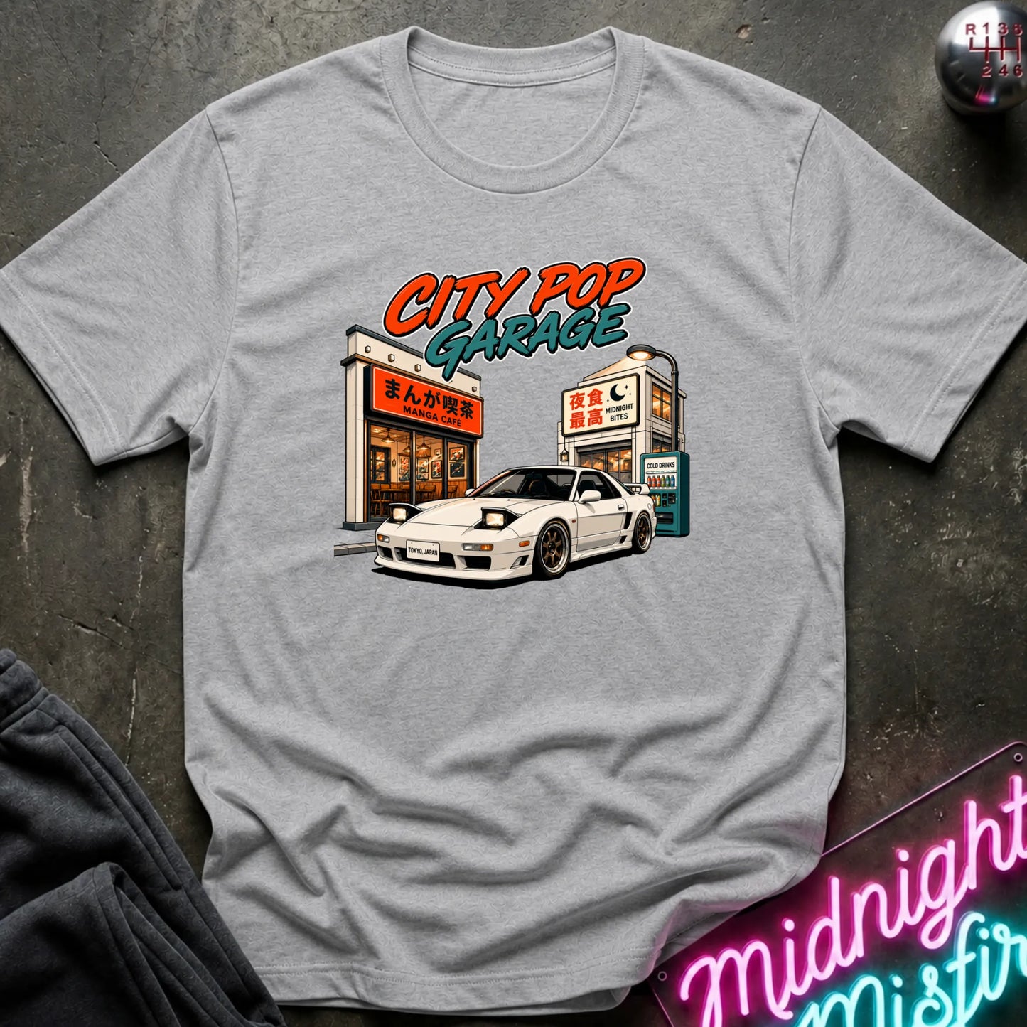 A light gray short-sleeve graphic t-shirt laid flat on a dark textured surface. The graphic features a white sports car parked in front of a cafe with the text 'CITY POP GARAGE' in orange and teal.
