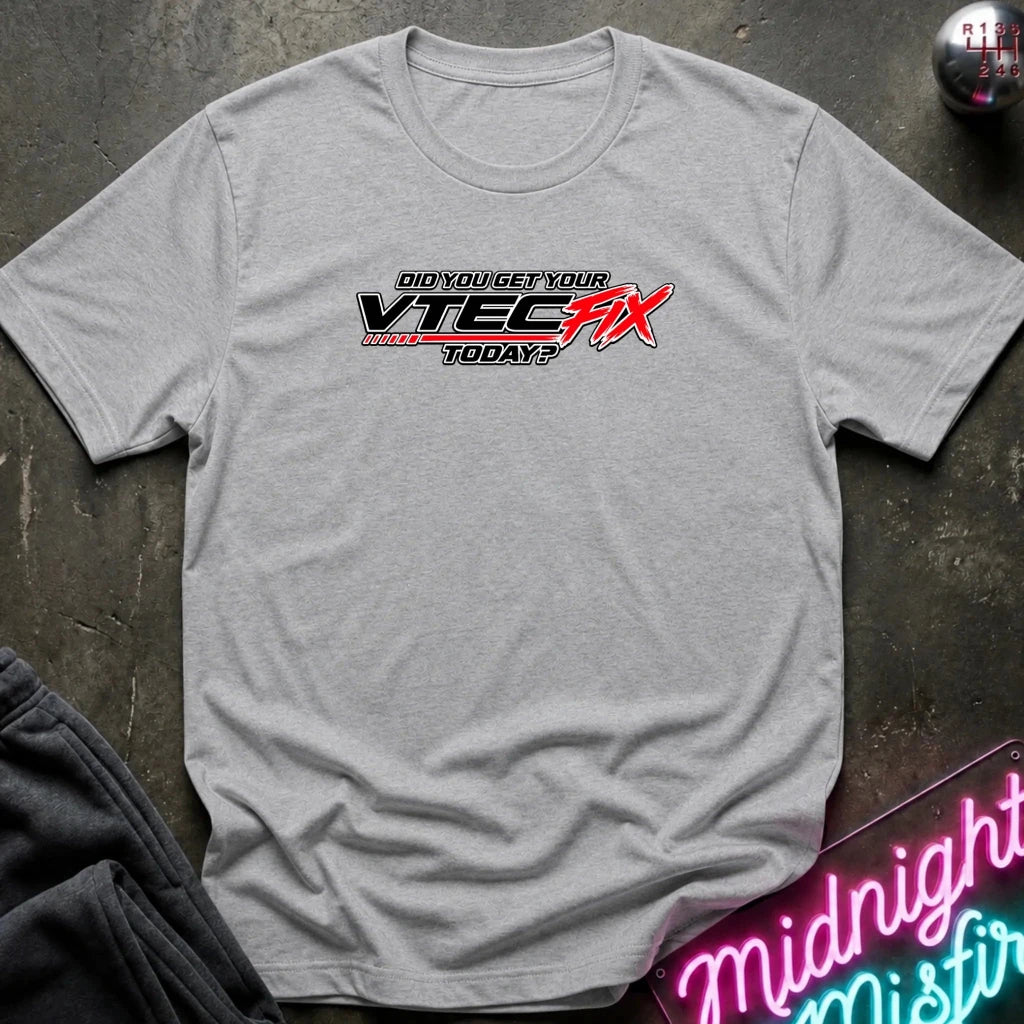 A heather gray short-sleeve t-shirt laid flat on a dark textured surface, featuring a red and black graphic print on the chest that reads 'Did you get your VTEC fix today?'. A neon sign and folded dark fabric are partially visible around the shirt.