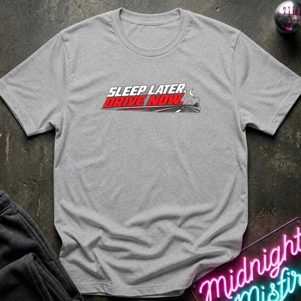 A heather gray short-sleeve t-shirt laid flat on a dark textured surface, featuring a chest graphic that reads 'SLEEP LATER DRIVE NOW' in white and red lettering with a small train illustration. 