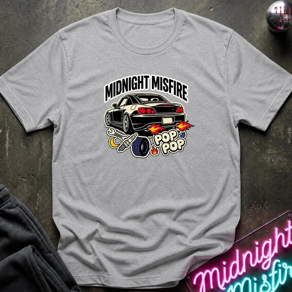 A light gray crewneck t-shirt featuring a graphic print of a black sports car with flames, the text 'MIDNIGHT MISFIRE', and 'POP POP' in a neon-style design, laid flat on a dark textured surface.