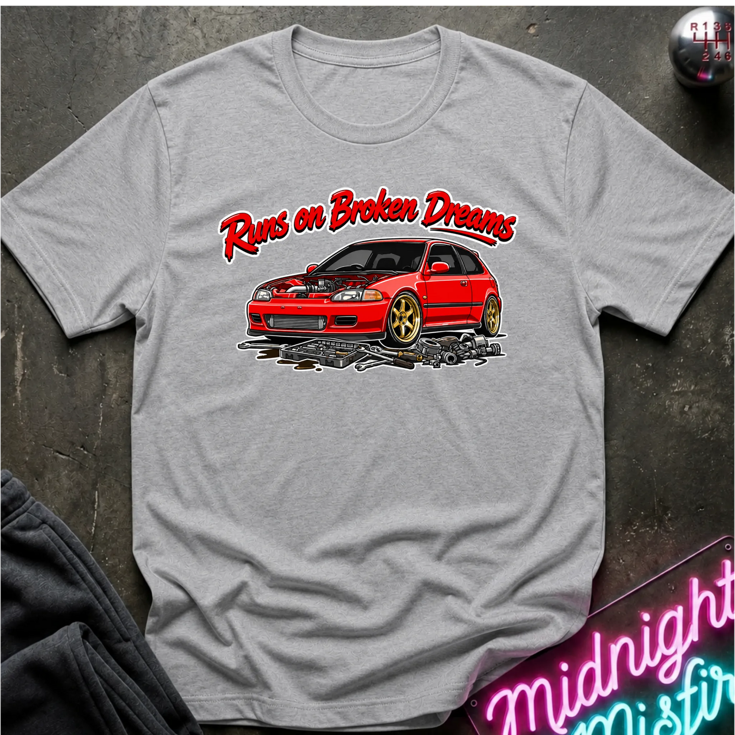 A light gray graphic t-shirt laid flat on a dark textured surface. The shirt features a red sports car with gold wheels and the text 'Runs on Broken Dreams' in red and white.