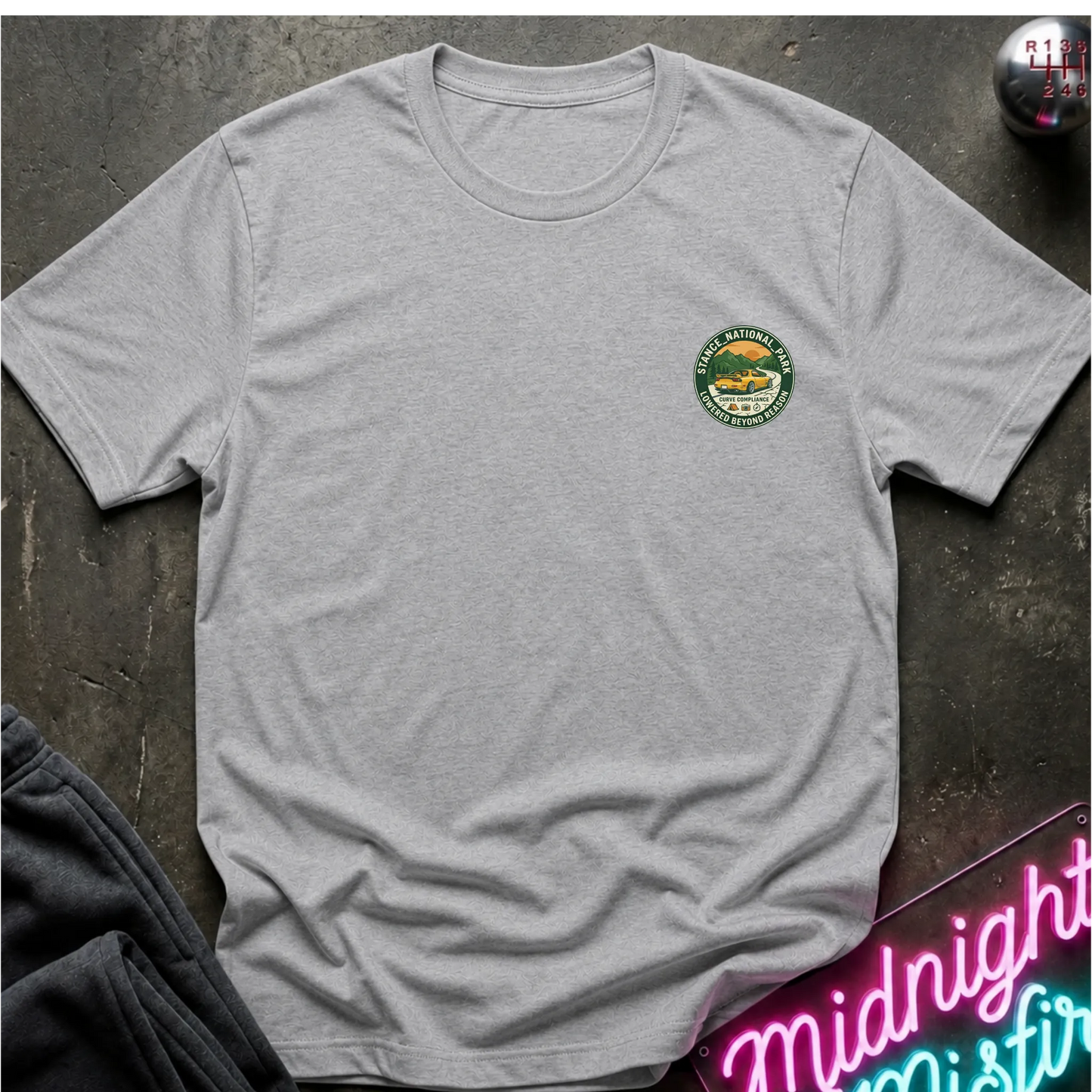 A light gray  crewneck t-shirt with a circular logo on the left chest, laid flat on a dark textured surface with a neon sign and dark pants in the background.