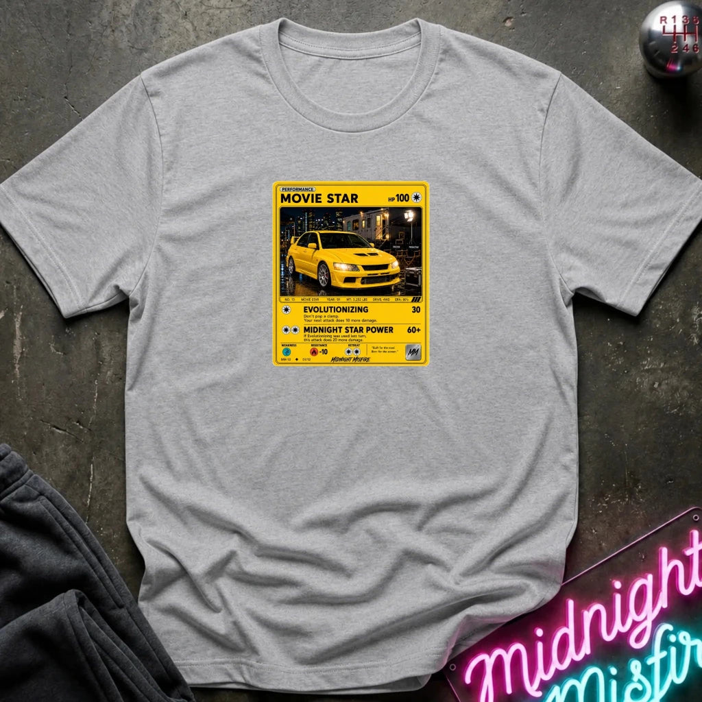 grey short-sleeve t-shirt laid flat on a dark textured surface, featuring a yellow and black retro-style graphic on the chest with a yellow car photo and text including 'MOVIE STAR'. 