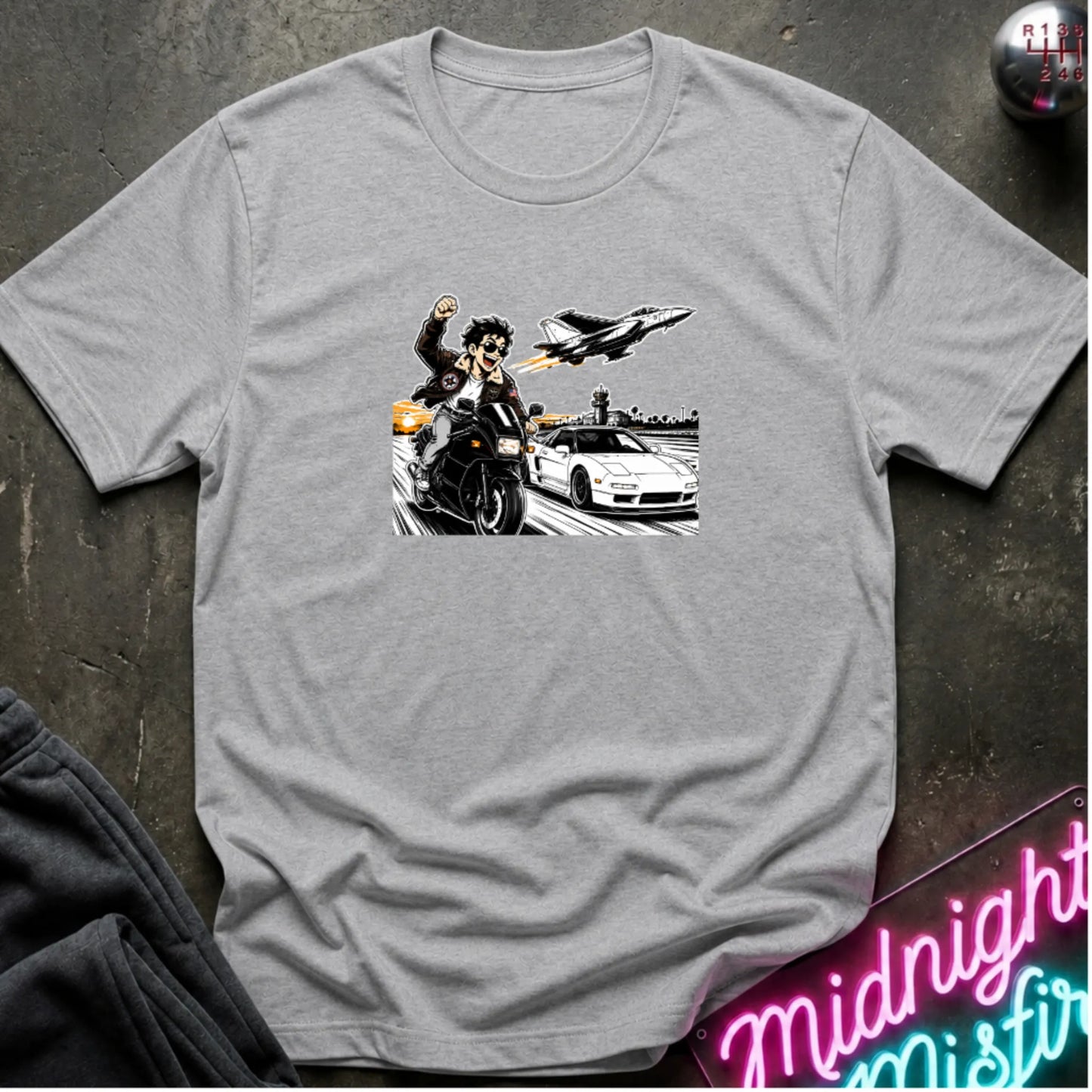 A heather gray short-sleeve crewneck t-shirt with a graphic print of a motorcycle racer, a sports car, and a jet plane, laid flat on a dark textured background with a neon sign and sweatpants partially visible.