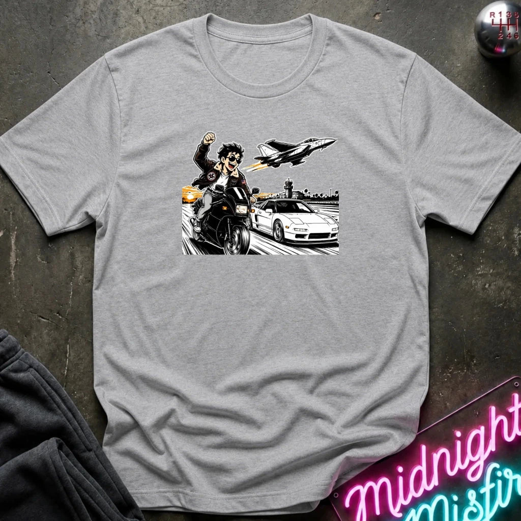 A light gray heather crewneck t-shirt featuring a graphic print of a motorcycle racer, a sports car, and a jet plane, laid flat on a dark textured surface with a neon sign and dark pants partially visible.