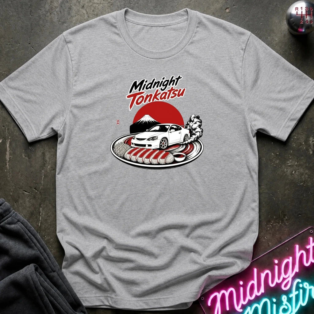A heather gray short-sleeve t-shirt laid flat on a dark textured surface. The shirt features a graphic print in the center with the words 'Midnight Tonkatsu' above an illustration of a white sports car, a red sun, and a black mountain.