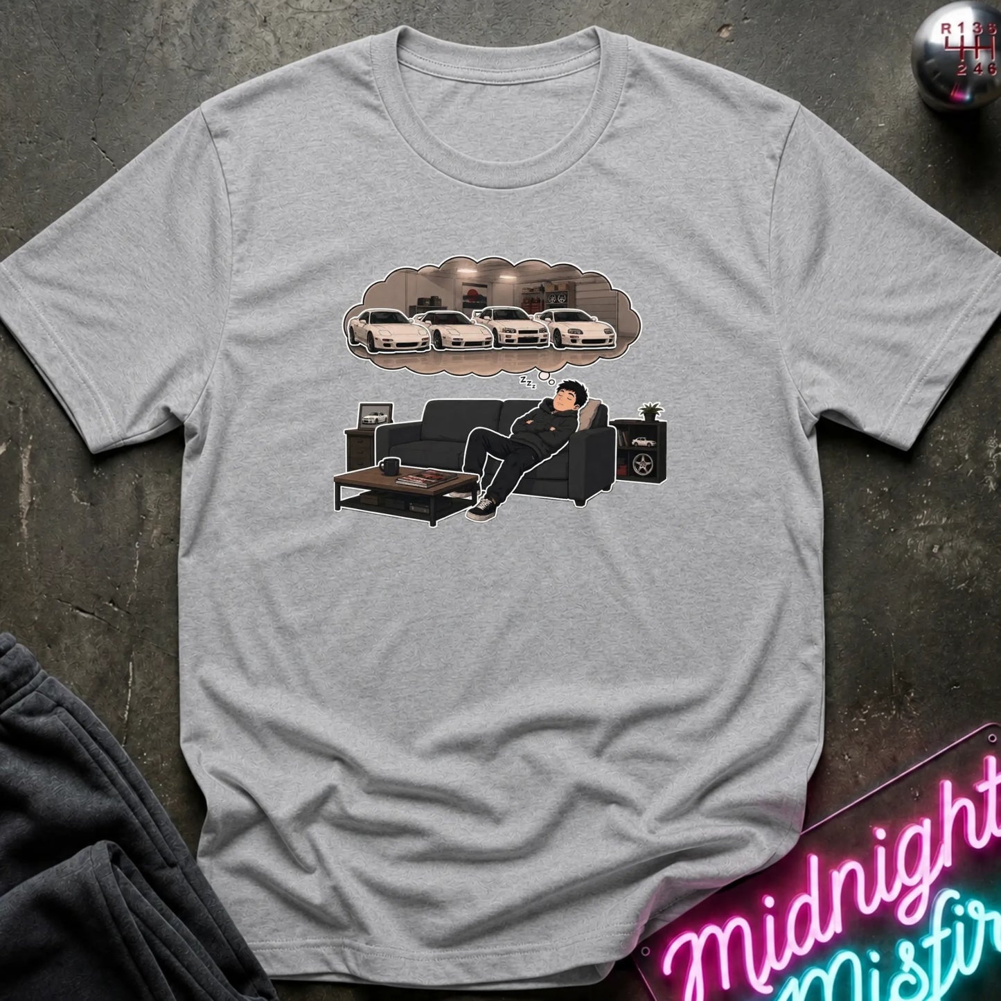 A light gray heather crewneck t-shirt laid flat on a dark textured surface. The shirt features a graphic print of a man relaxing on a black couch with a coffee table, with a thought bubble above showing three customized white cars.