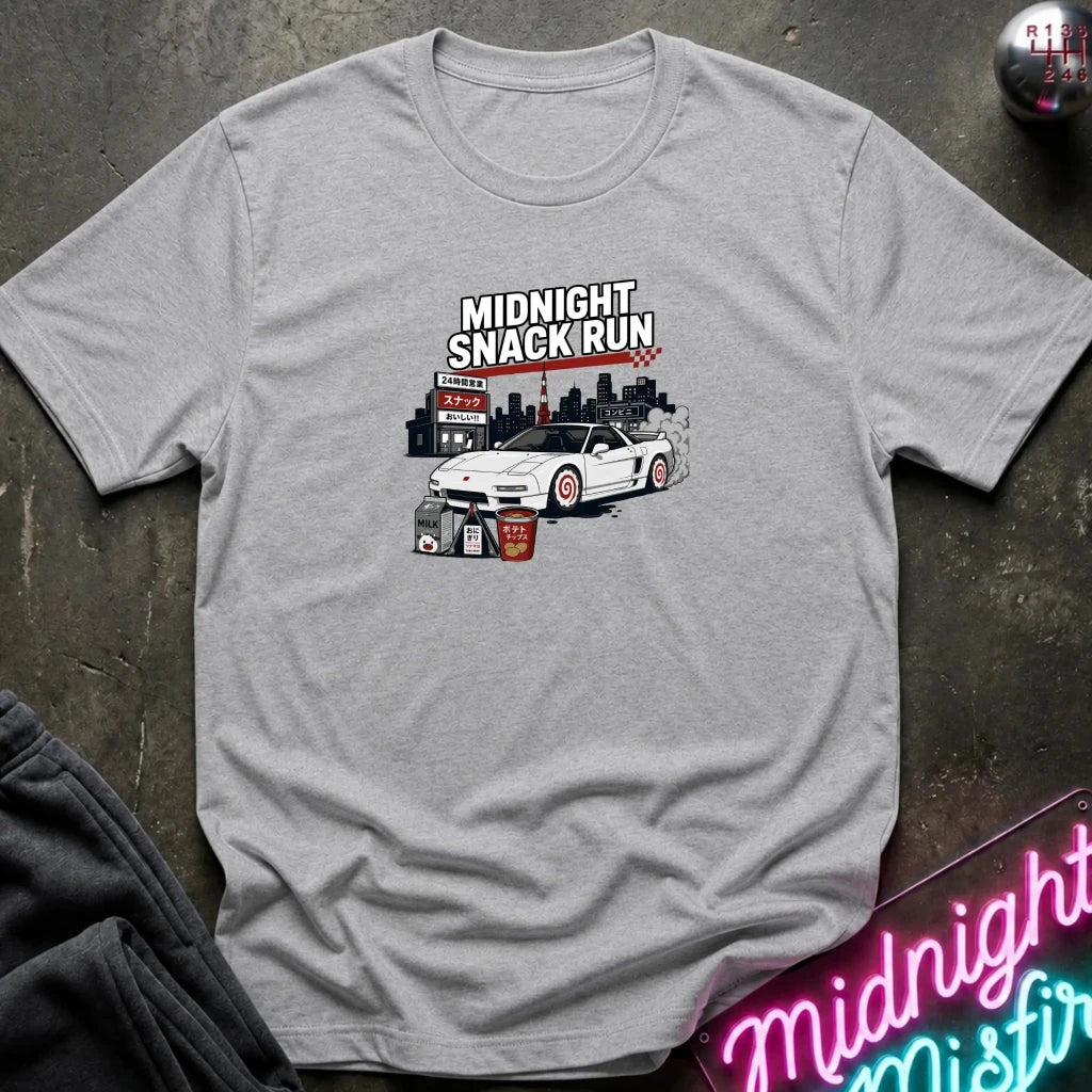 A light gray heather crewneck t-shirt featuring a graphic print of a white sports car with a checkered flag and the text 'MIDNIGHT SNACK RUN' above it. The shirt is laid flat on a dark textured surface with a neon sign and dark sweatpants partially visible.