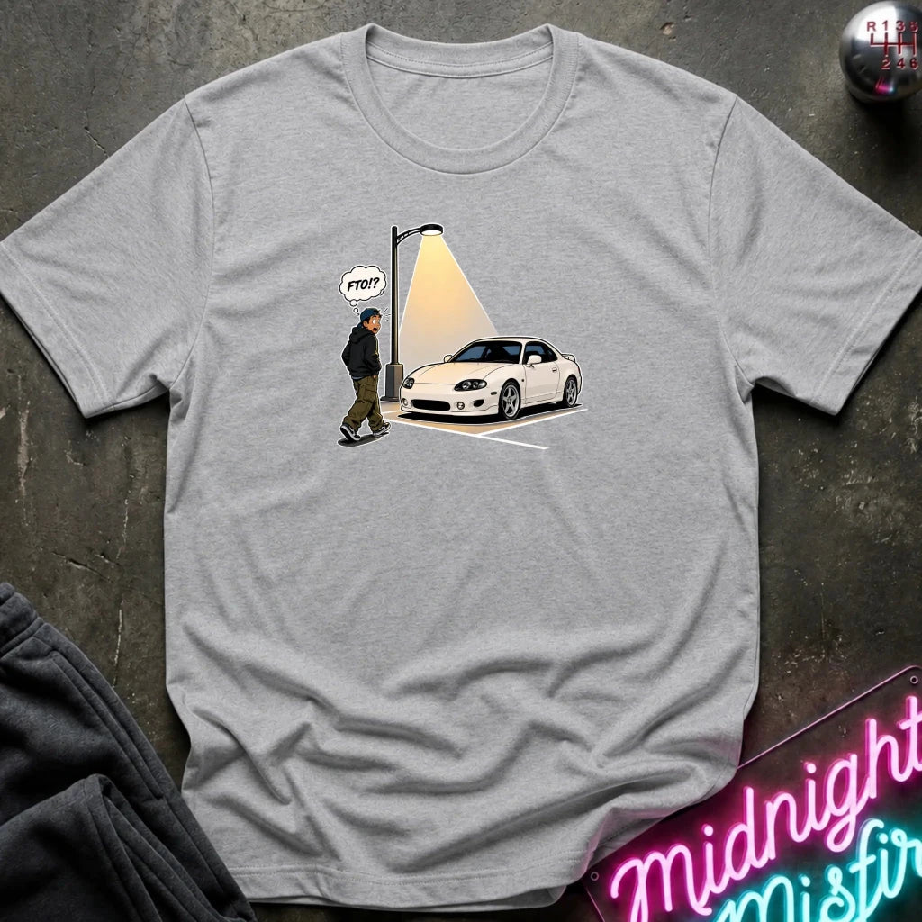 A heather gray short-sleeve t-shirt laid flat on a dark textured surface, featuring a graphic print of a white sports car parked under a streetlamp with a walking figure and a speech bubble 
