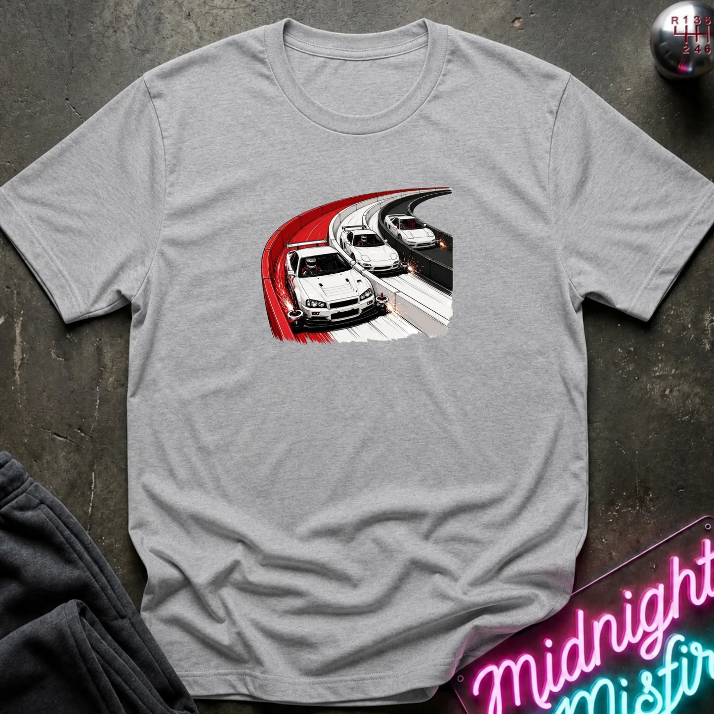 A light gray heather crewneck t-shirt featuring a graphic print of three white sports cars racing on a highway with a red curve, laid flat on a dark textured surface.