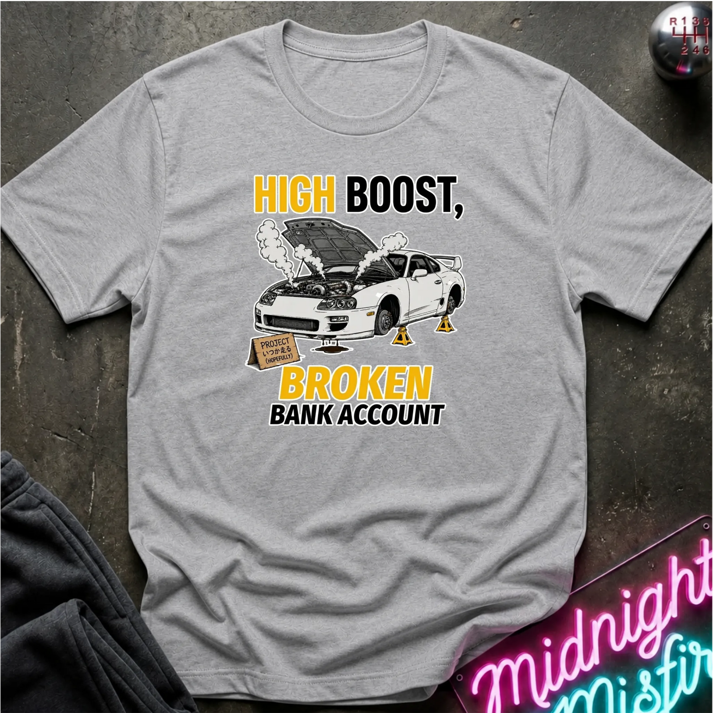 A light gray crewneck t-shirt with a humorous graphic print. The design features a white sports car with its hood open, emitting smoke, with yellow and black text reading 'HIGH BOOST, BROKEN BANK ACCOUNT'.
