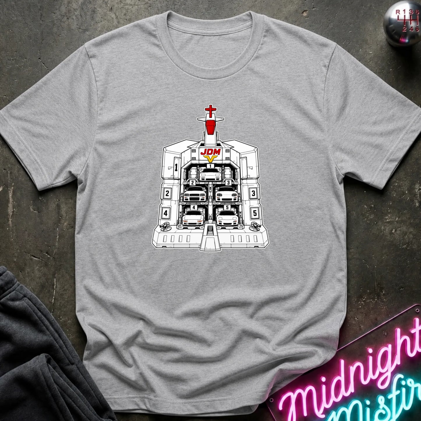 A light gray short-sleeve crewneck t-shirt laid flat on a dark textured surface, featuring a black and white graphic of the JDM (Japanese Domestic Market) cars in a launch bay.