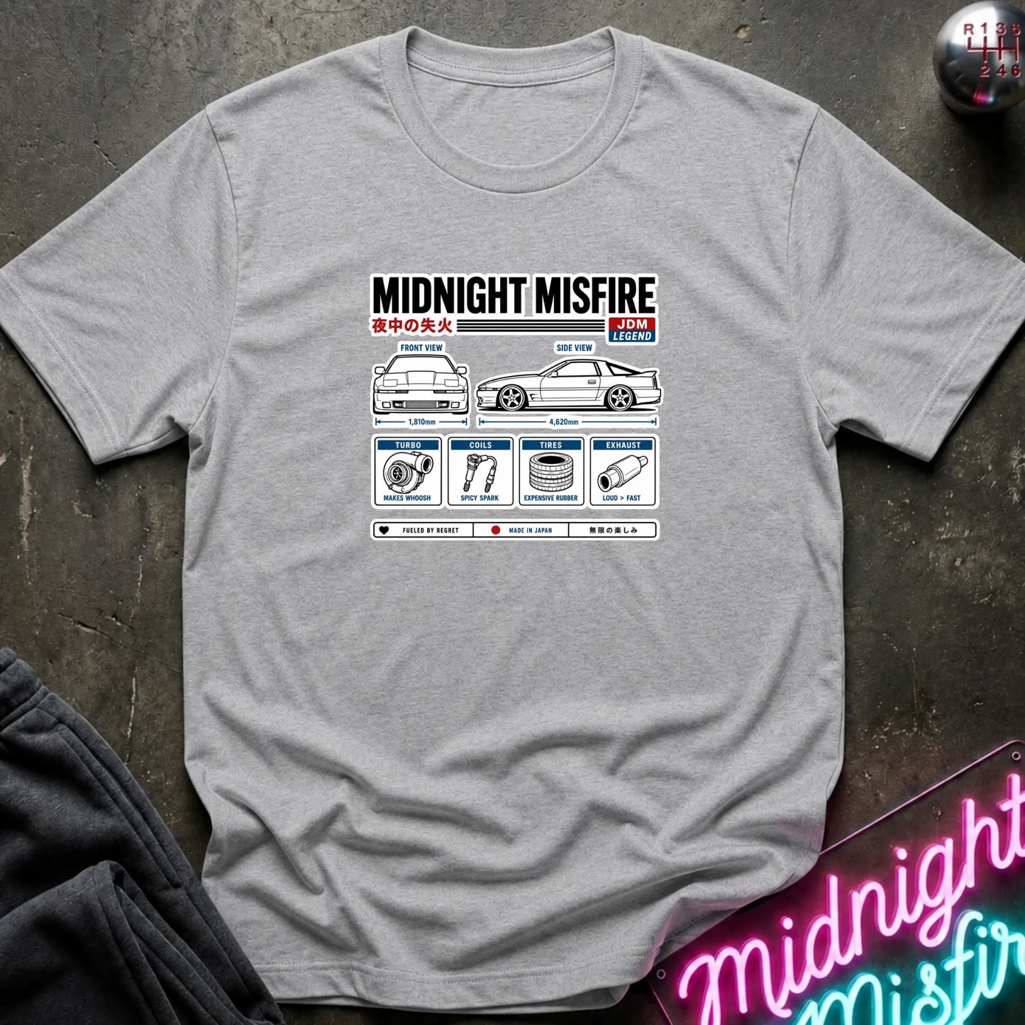 A light gray heather crewneck t-shirt featuring a 'Midnight Misfire' car diagnostic graphic print on the chest, laid flat on a dark textured surface with a neon sign in the corner.