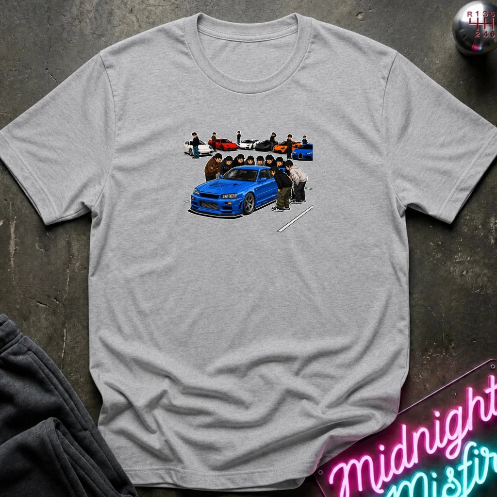 grey short-sleeve crewneck t-shirt laid flat on a dark textured surface. The center of the shirt features a graphic print of a blue sports car with a group of people standing behind it.