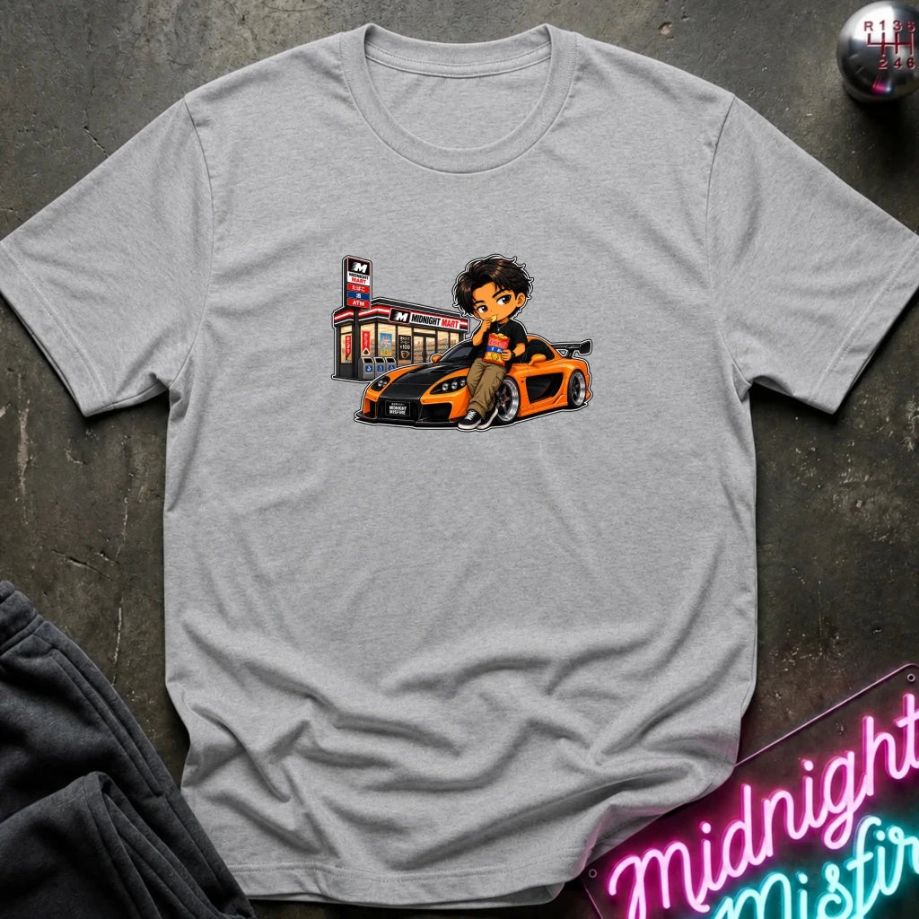A light heather gray short-sleeve crewneck t-shirt laid flat on a dark textured surface, featuring a cartoon graphic of a man sitting on a orange and black sports car next to a store.
