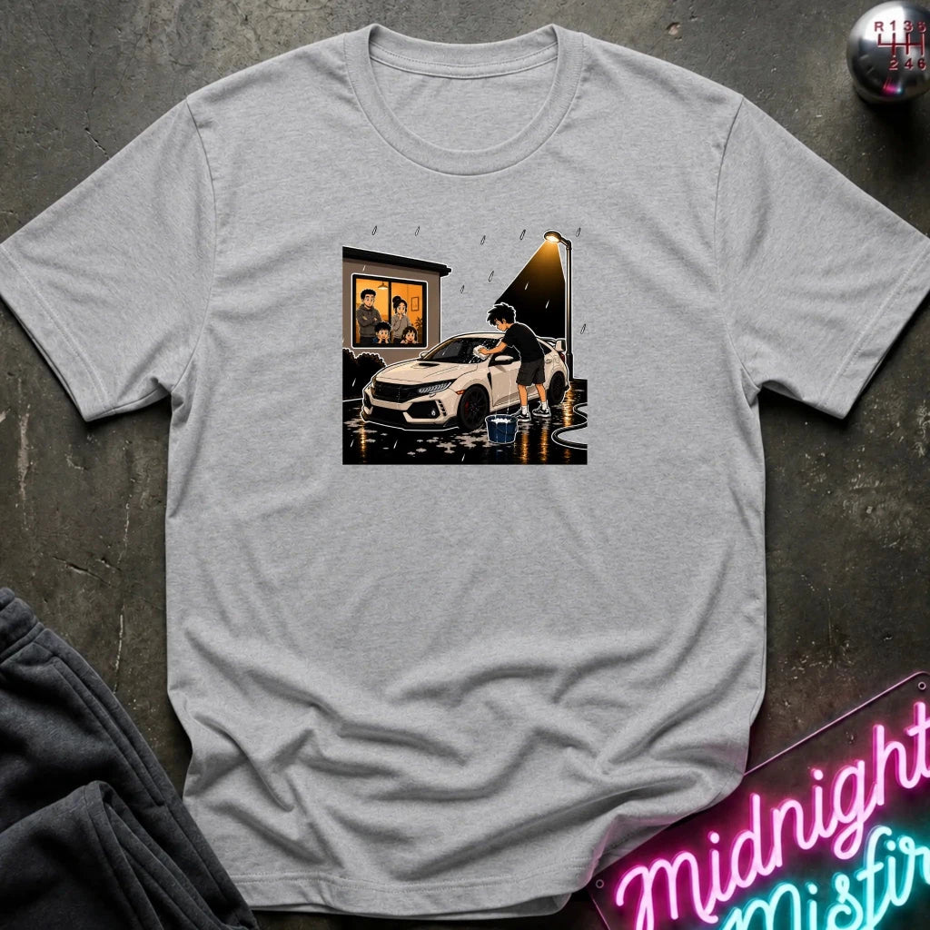 A light gray heather crewneck t-shirt laid flat on a dark textured surface. The center of the shirt features a rectangular graphic print depicting a white sports car in a rain-slicked garage scene with a person standing by the open door.