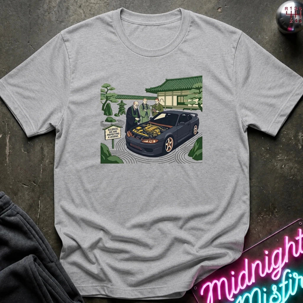 A light gray heather crewneck t-shirt featuring a graphic print of a dark blue car with its hood open, surrounded by people in a Japanese garden setting. The shirt is displayed flat on a dark textured surface with a neon sign and dark sweatpants partially visible.