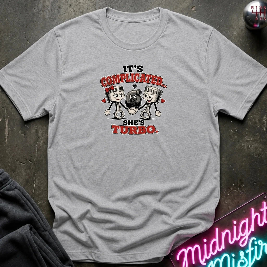 A light gray short-sleeve graphic T-shirt laid flat on a dark textured surface. The shirt features a cartoon graphic of two pistons holding a heart-shaped turbocharger, with the text 'IT'S COMPLICATED.