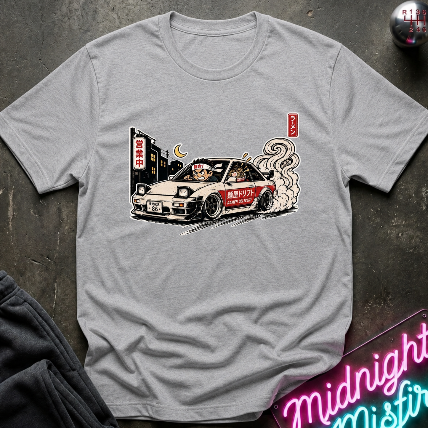 A light gray crewneck t-shirt featuring a graphic print of a white car with cartoon characters and smoke, laid flat on a dark textured surface.