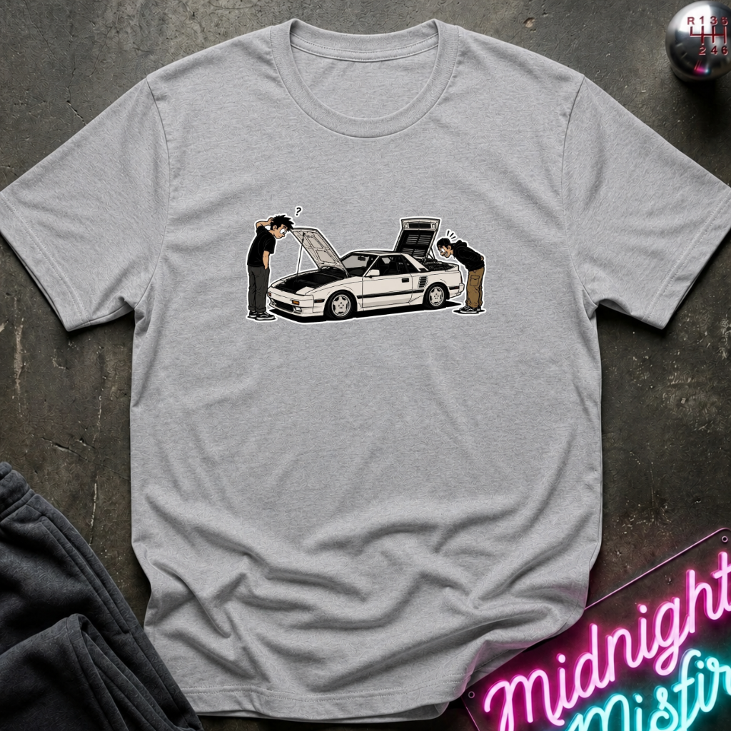 A light gray heather crewneck t-shirt laid flat on a dark textured surface. The shirt features a graphic print of a white sports car with its hood and trunk open, with two people leaning over it.