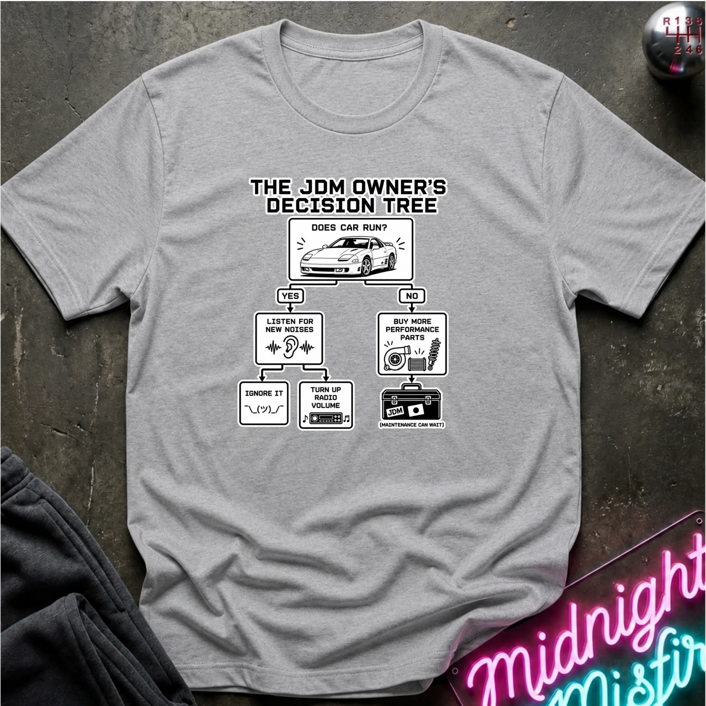 A heather gray short-sleeve graphic T-shirt laid flat on a dark textured surface. The shirt features a black printed decision tree titled 'THE JDM OWNER'S DECISION TREE' with illustrations and text related to car care and maintenance.