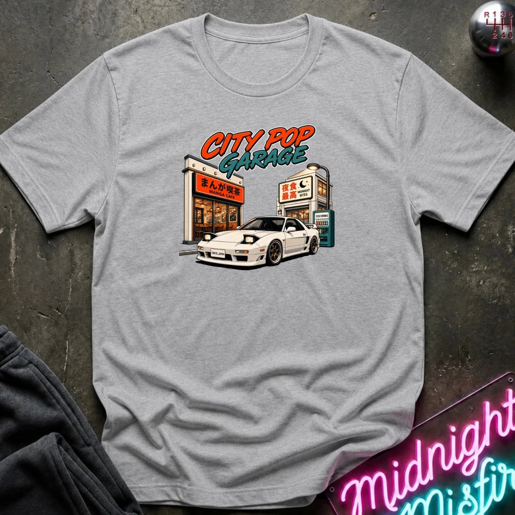 A light gray heather crewneck t-shirt featuring a retro-style graphic of a white sports car parked outside 'City Pop Garage' with orange and teal lettering, laid flat on a dark textured surface.