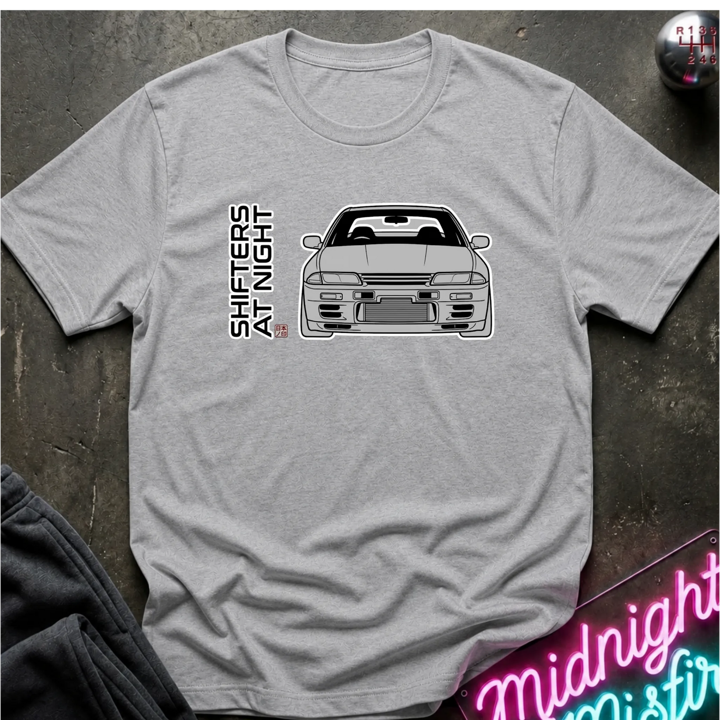A light gray heather crewneck t-shirt featuring a black line art graphic of a sports car and the vertical text 'SHIFTERS AT NIGHT' on the front, laid flat on a dark textured surface.