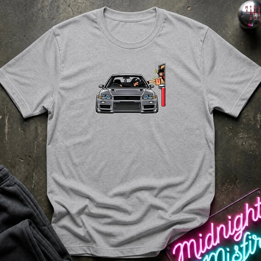 A light heather gray crewneck t-shirt laid flat on a dark textured surface. The center of the shirt features a graphic print of a gray sports car facing forward, with a red and white striped pole or sign on the right side of the car.