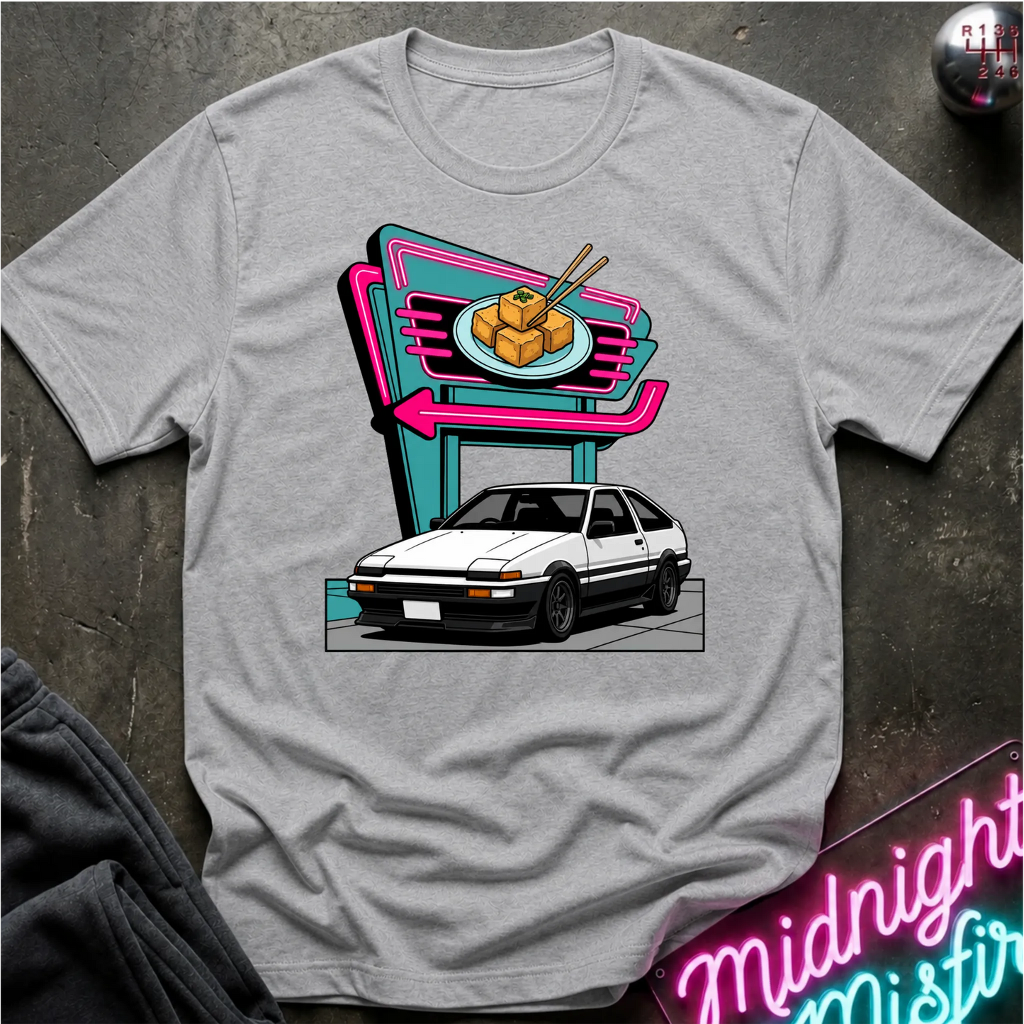 A heather gray short-sleeve crewneck t-shirt featuring a retro-style graphic of a white sports car parked in front of a neon sign. The shirt is displayed flat on a dark textured surface with a dark sweatshirt and a neon sign partially visible as props.