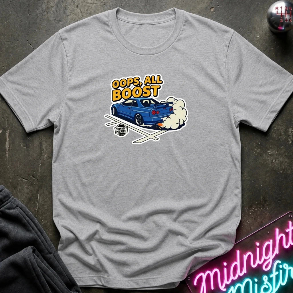 A heather gray crewneck t-shirt featuring a graphic of a blue sports car with smoke and the text 'OOPS, ALL BOOST' in yellow and orange. The shirt is laid flat on a dark textured surface with a neon sign and dark pants partially visible.