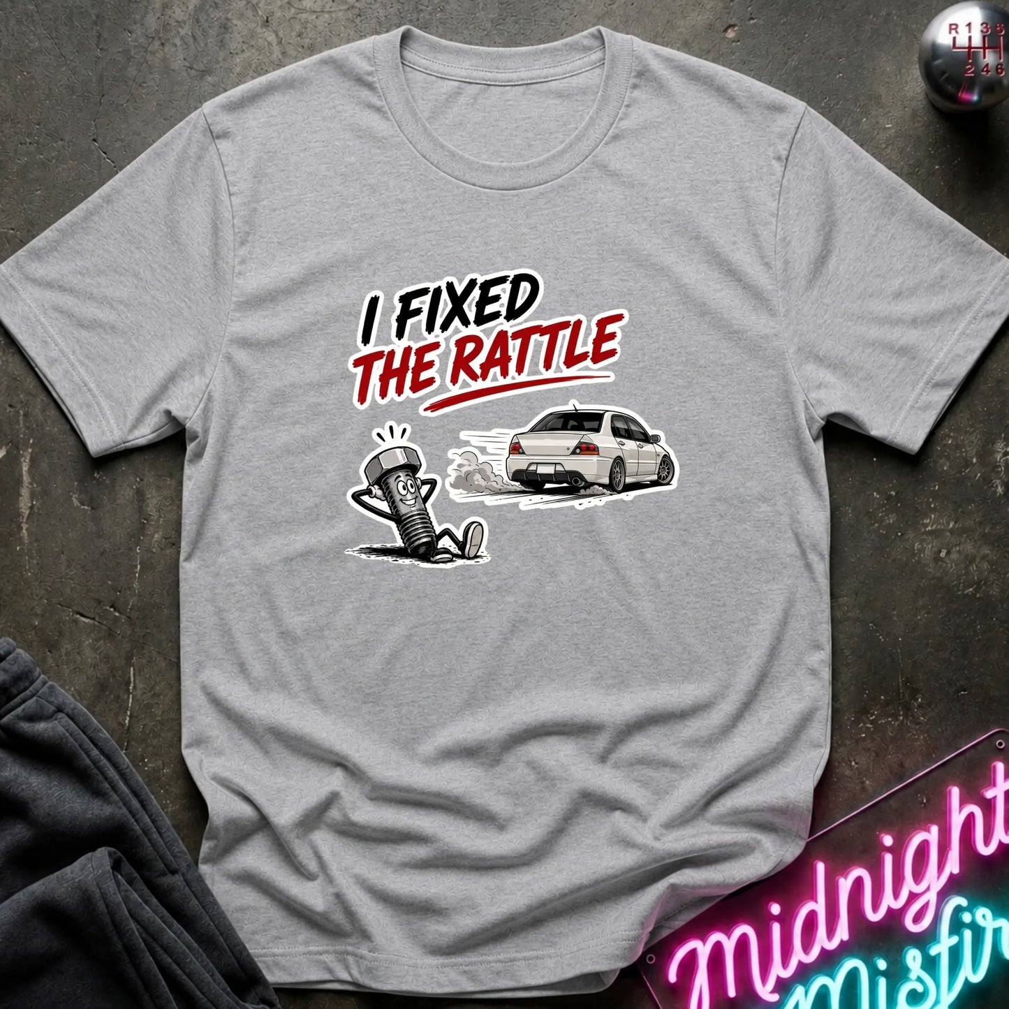 A light gray heather crewneck t-shirt laid flat on a dark textured surface. The shirt features a graphic print with the text 'I FIXED THE RATTLE' in black and red, above an illustration of a white car and a cartoon figure.